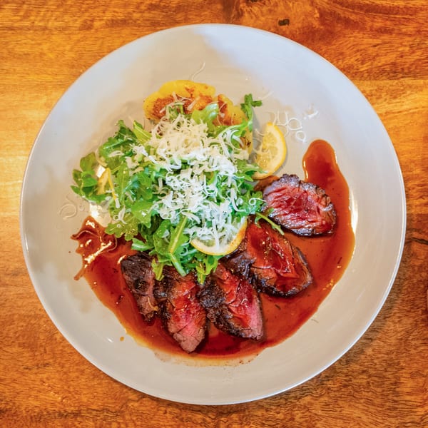 Seared steak slices with a rich sauce, topped with arugula, shredded cheese, and lemon on a white plate.