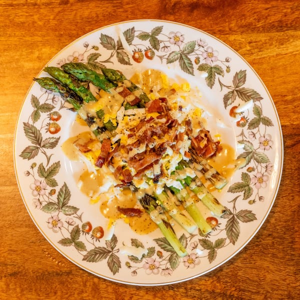 Grilled asparagus spears topped with bacon, chopped egg, and creamy dressing on a floral plate.
