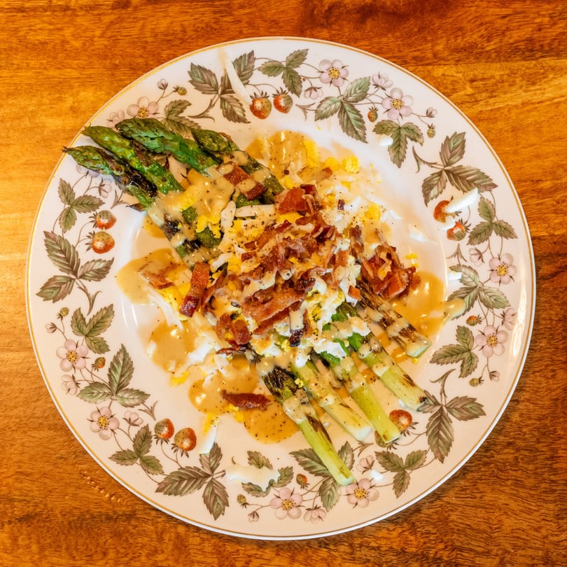 Grilled asparagus spears topped with bacon, chopped egg, and creamy dressing on a floral plate.