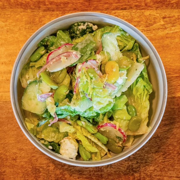 Overhead view of a fresh green salad with lettuce, radishes, cucumbers, and edamame in a white bowl.