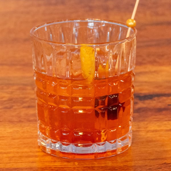 An Old Fashioned cocktail in a textured glass, garnished with an orange peel and a cherry.