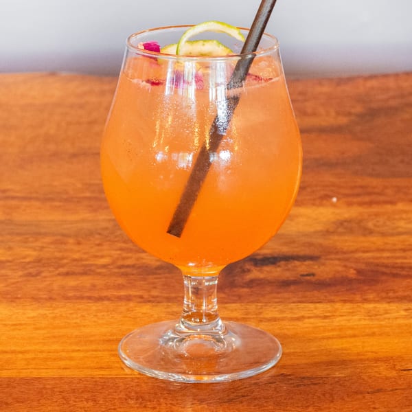 A vibrant orange cocktail in a stemmed glass, garnished with lime peel and rose petals, on a wooden table.