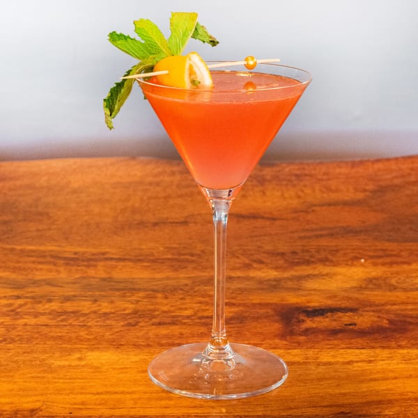 A vibrant red cocktail in a martini glass, garnished with mint and a citrus slice, on a wooden table.