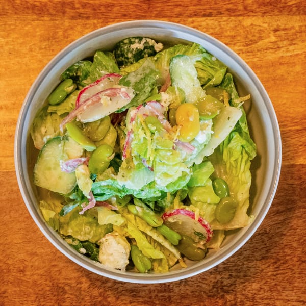 Overhead view of a fresh green salad with radishes, edamame, and creamy dressing in a white bowl.